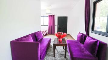 A room with purple sofas, a wooden table, and a window with curtains.