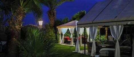 A tented outdoor area with a lawn, palm trees, and a table with chairs.