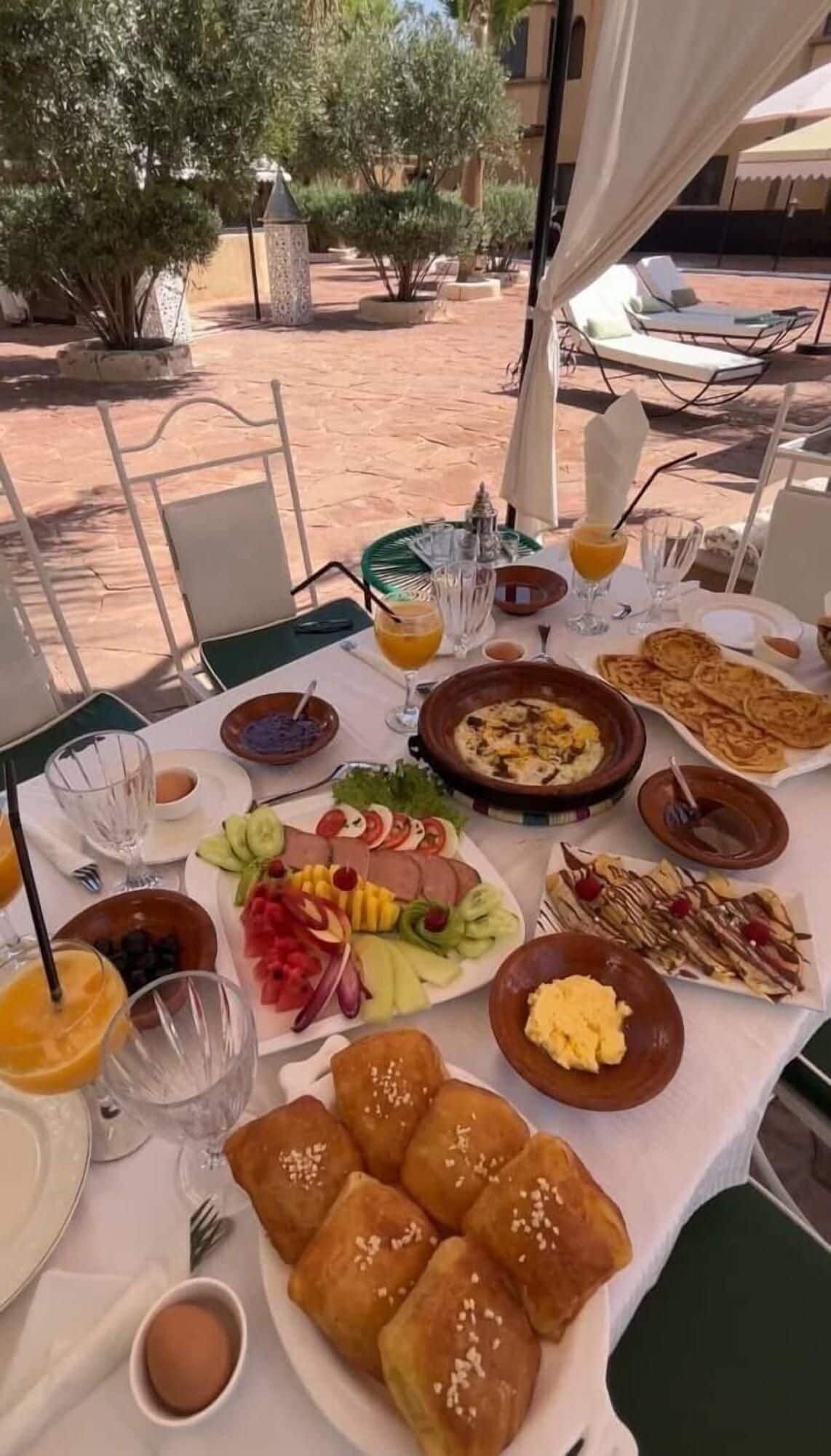 An outdoor dining table with various dishes, including fruits, bread, and a hot dish, set against a backdrop of a building and trees.