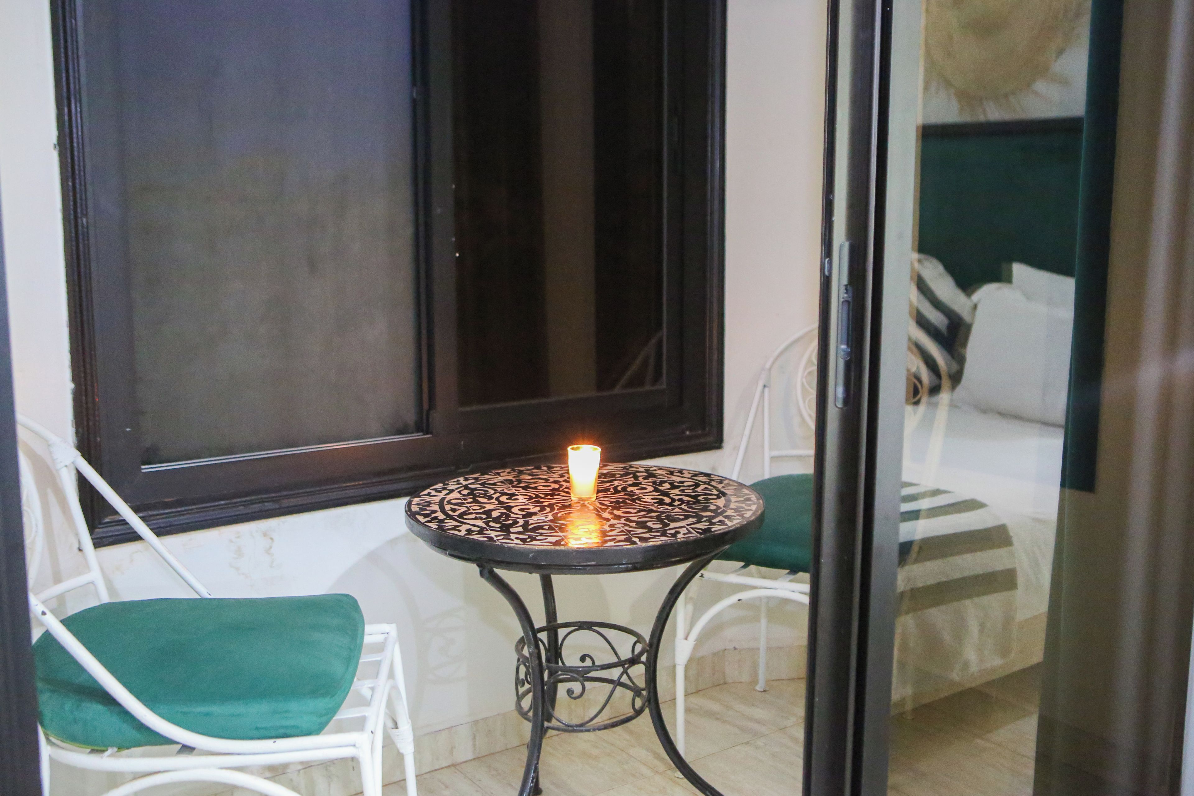 A small outdoor seating area with a table and two chairs, a lit candle, and a window with a view of a bed.