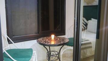 A small outdoor seating area with a table and two chairs, a lit candle, and a window with a view of a bed.