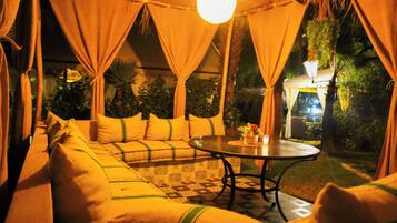 A cozy outdoor seating area with a sofa and a round table, surrounded by curtains and a hanging light fixture.