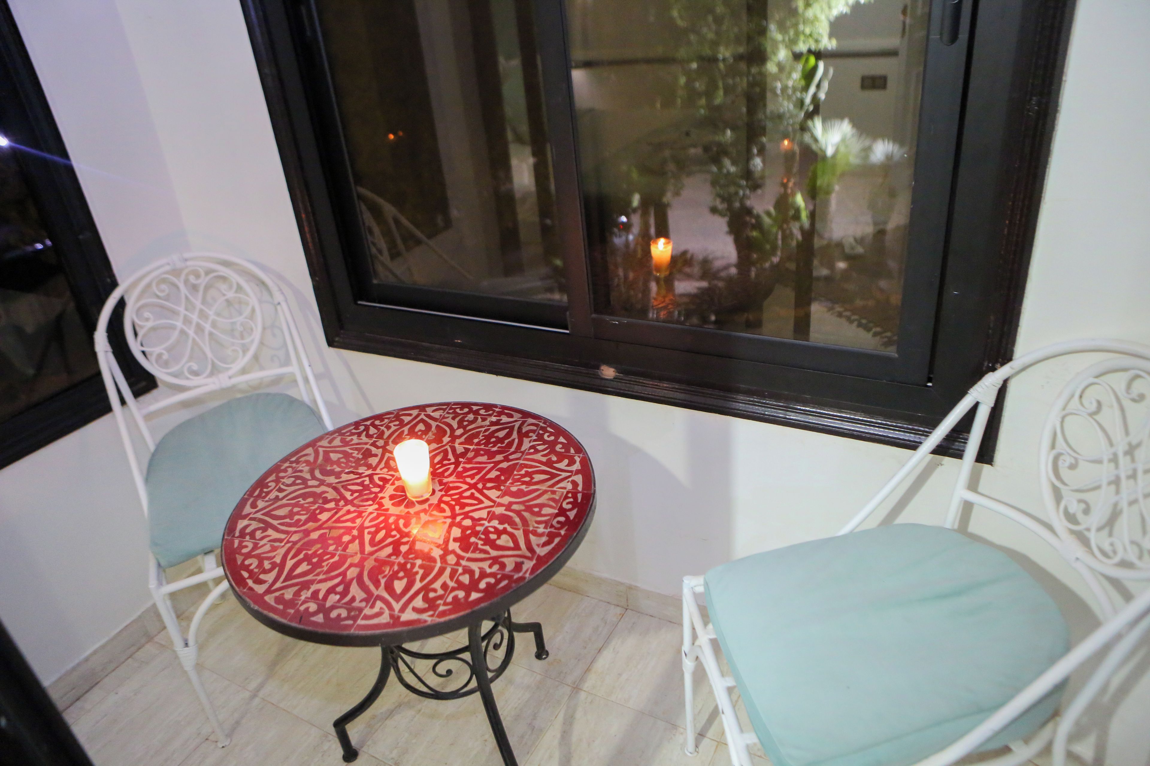 Ein kleiner Balkon mit Tisch und zwei Stühlen, eine Kerze auf dem Tisch und Blick auf einen Garten durch ein Fenster.