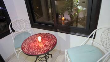 Ein kleiner Balkon mit Tisch und zwei Stühlen, eine Kerze auf dem Tisch und Blick auf einen Garten durch ein Fenster.