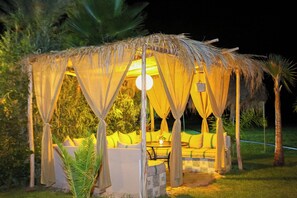 A thatched-roof outdoor seating area with cushions and a table, surrounded by greenery. - Weedan Ferme D'hôtes (Marrakech)