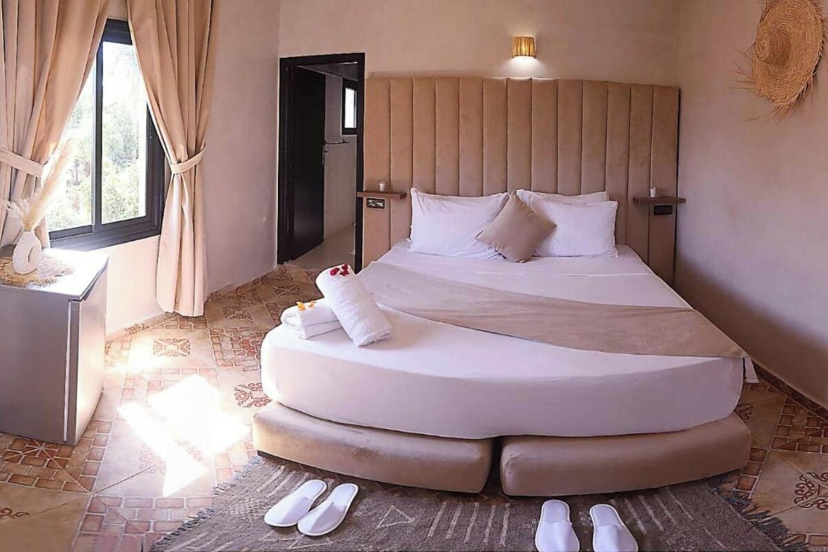 A spacious bedroom with a curved bed, a bedside table, and a large window with curtains.