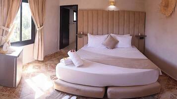 A spacious bedroom with a curved bed, a bedside table, and a large window with curtains.