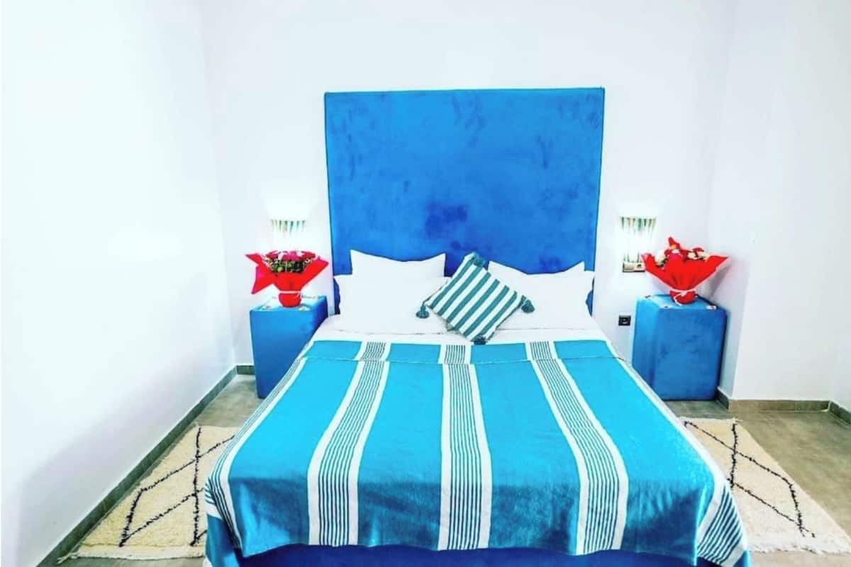 Una cama con cabecera azul, almohadas blancas y ropa de cama a rayas. Hay dos mesitas de noche con lámparas rojas y blancas.
