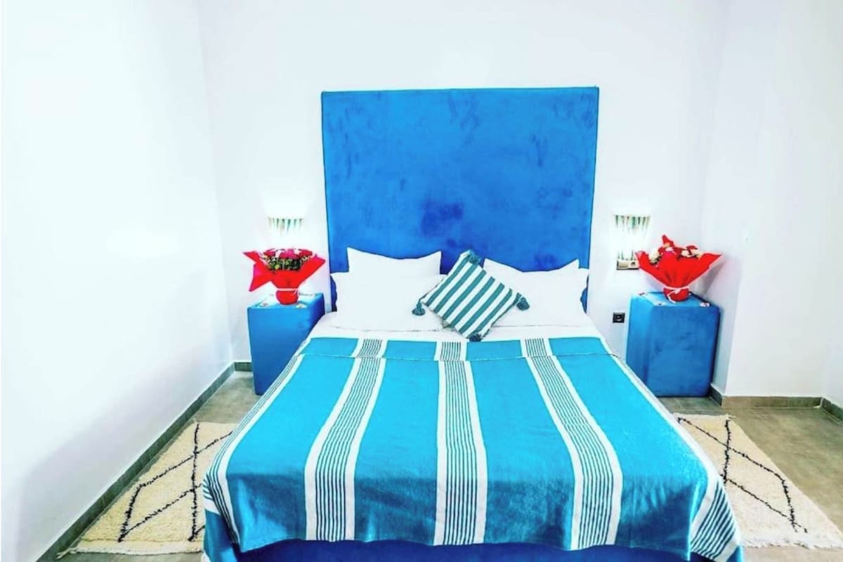 A bed with a blue headboard, white pillows, and striped bedding. There are two nightstands with red and white lamps.