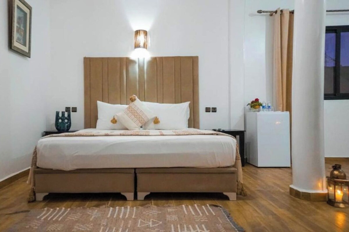 A bedroom with a bed, bedside tables, a nightstand, a wall-mounted light, a small refrigerator, and a window with curtains.