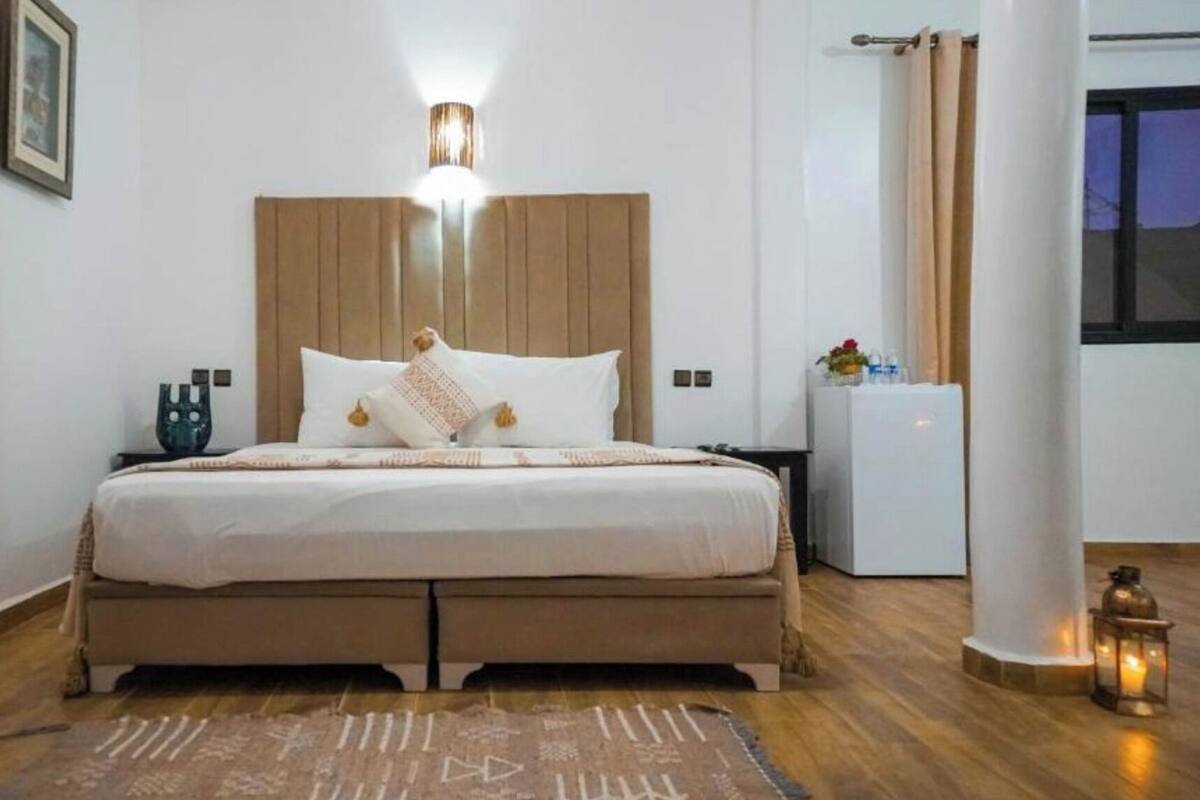 Un dormitorio con cama, mesitas de noche, refrigerador pequeño y ventana con cortinas.