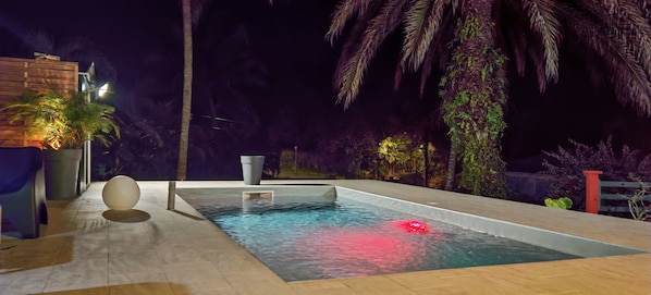 A rectangular swimming pool with a light-colored deck, surrounded by palm trees and plants, and a wooden fence in the background.