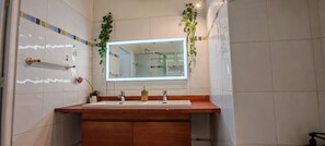 Une salle de bain avec un meuble-lavabo en bois, deux vasques, un miroir et des plantes vertes.