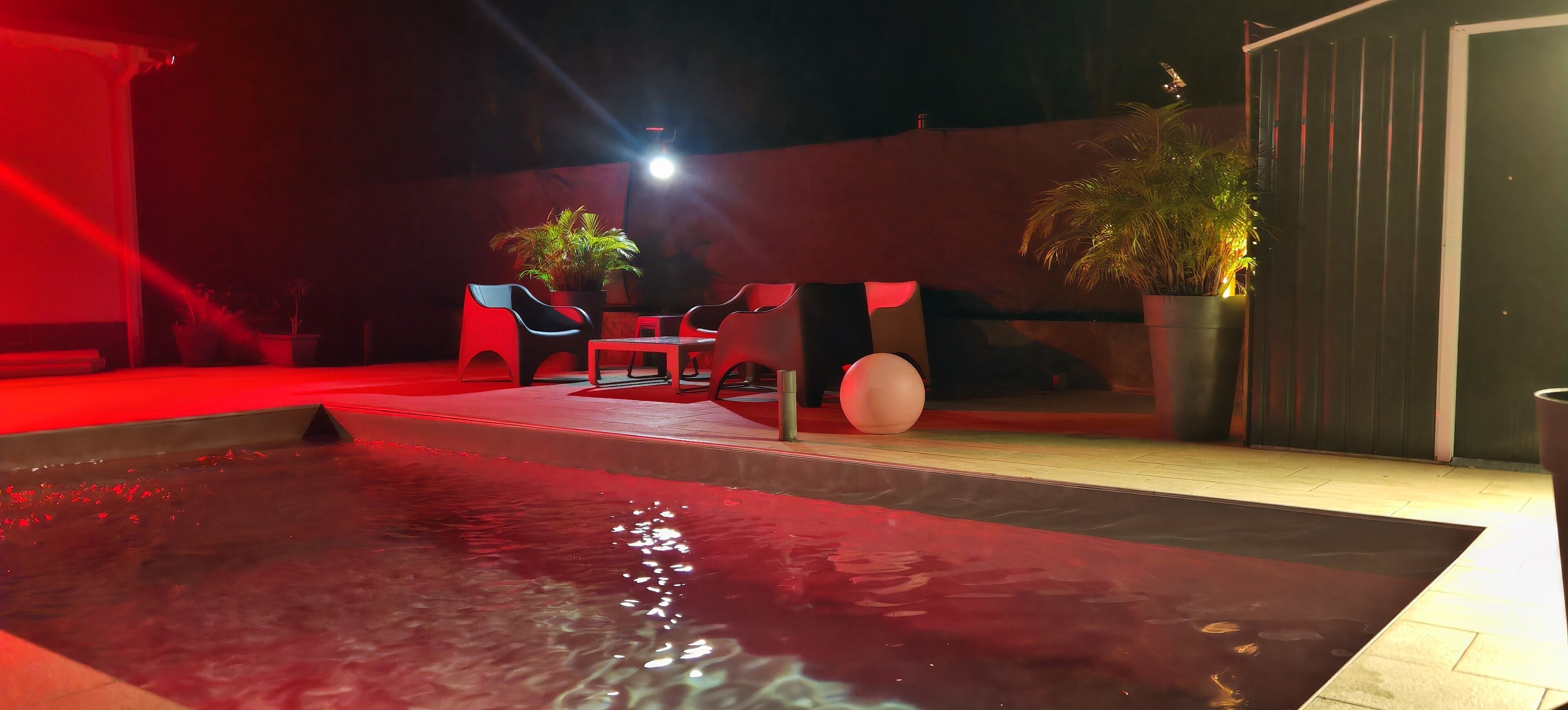 Un espace extérieur avec piscine, éclairé de lumières rouges, comprenant des sièges et des plantes en pot.