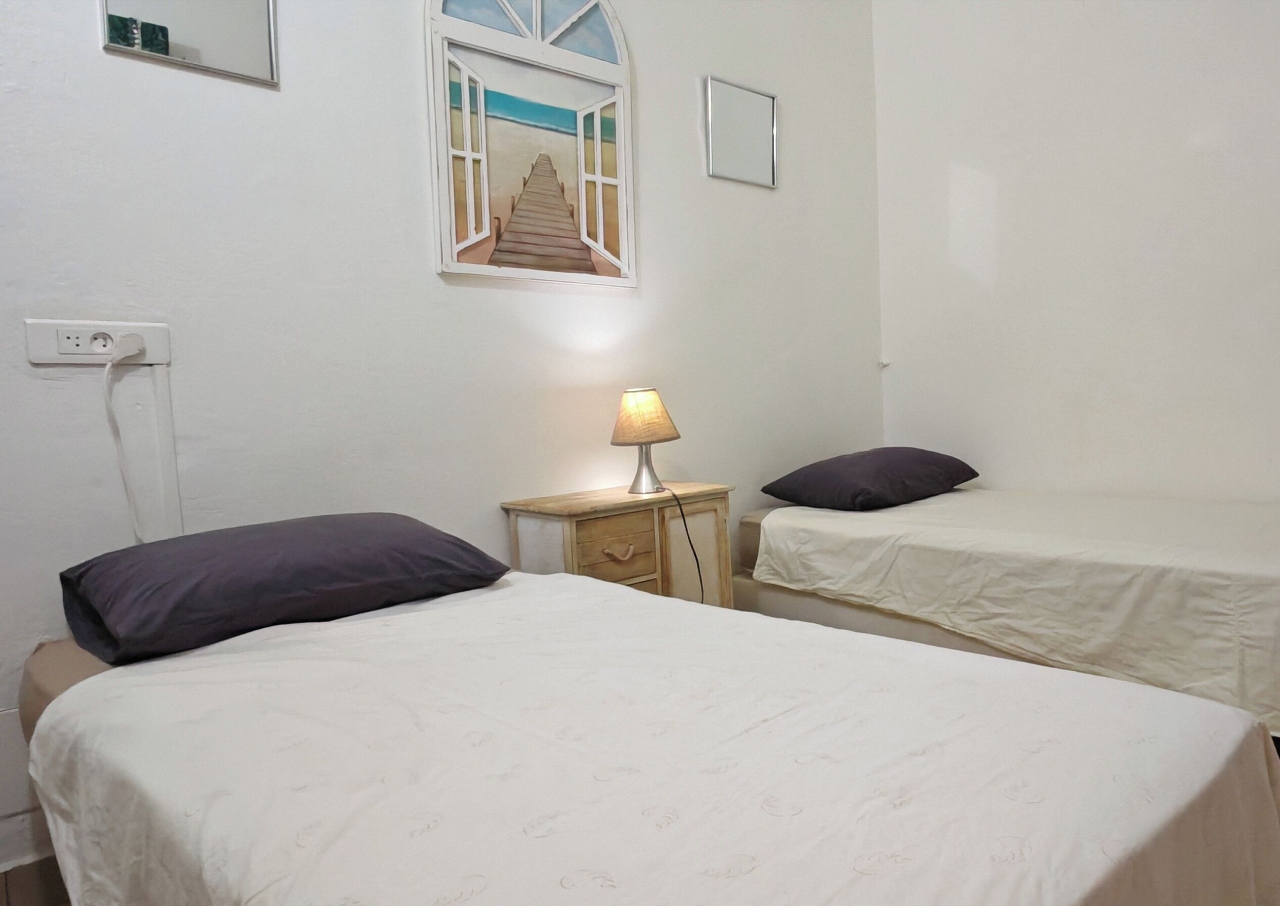 Ein Zimmer mit zwei Betten, einem Nachttisch mit Lampe, einer Steckdose und einem Fenster mit Blick auf Strand und Steg.