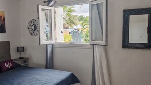 Un dormitorio con cama, mesita de noche, lámpara, pintura de paisaje urbano, espejo decorativo y ventana con cortinas.