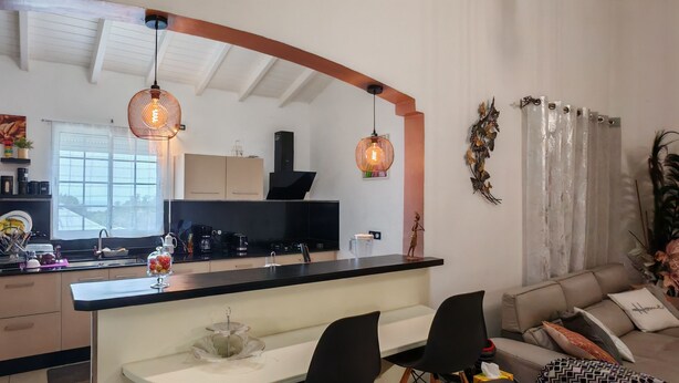 Una cocina moderna con una isla central, encimeras negras e iluminación colgante. Hay un comedor con un sofá y sillas.