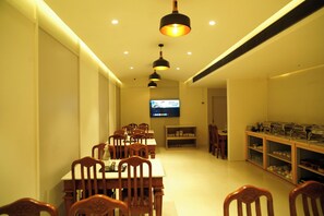 A dining area with wooden tables and chairs, a television mounted on the wall, and shelves with dishes and glasses. - Hotel Harriet (Rameshwaram)