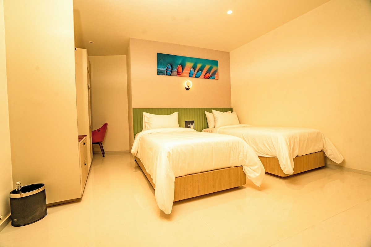 A hotel room with two beds, a painting on the wall, and a small red chair.