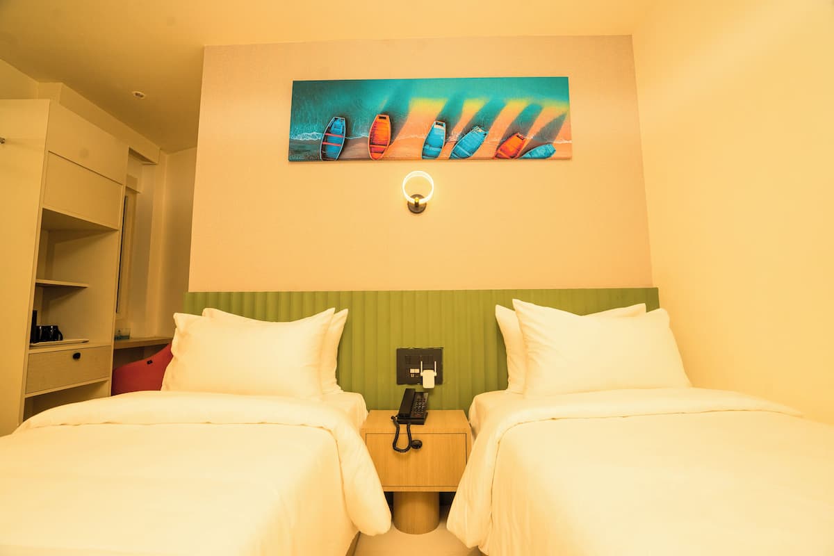 A hotel room with two beds, a painting of boats on the wall, a bedside table with a phone, and a green headboard.