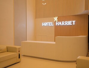 A hotel reception desk with a modern design, a sign for 'HOTEL HARRIET', and a plush sofa in the foreground. - Hotel Harriet (Rameshwaram)