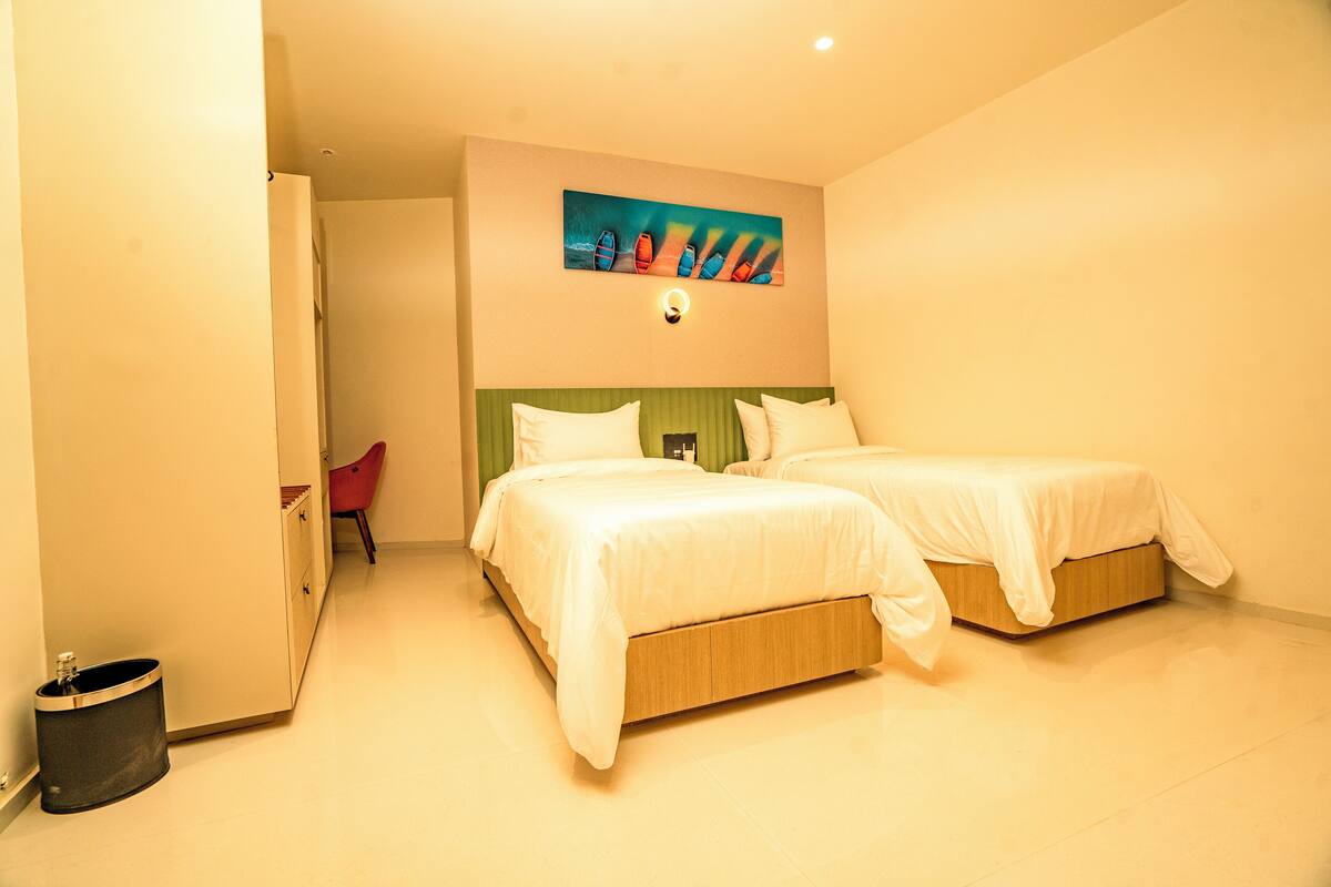 A hotel room with two beds, a painting on the wall, and a small red chair.
