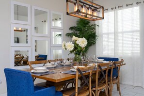Un comedor con una mesa de madera dispuesta para seis personas, rodeada de sillas azules, un espejo grande y una lámpara colgante.