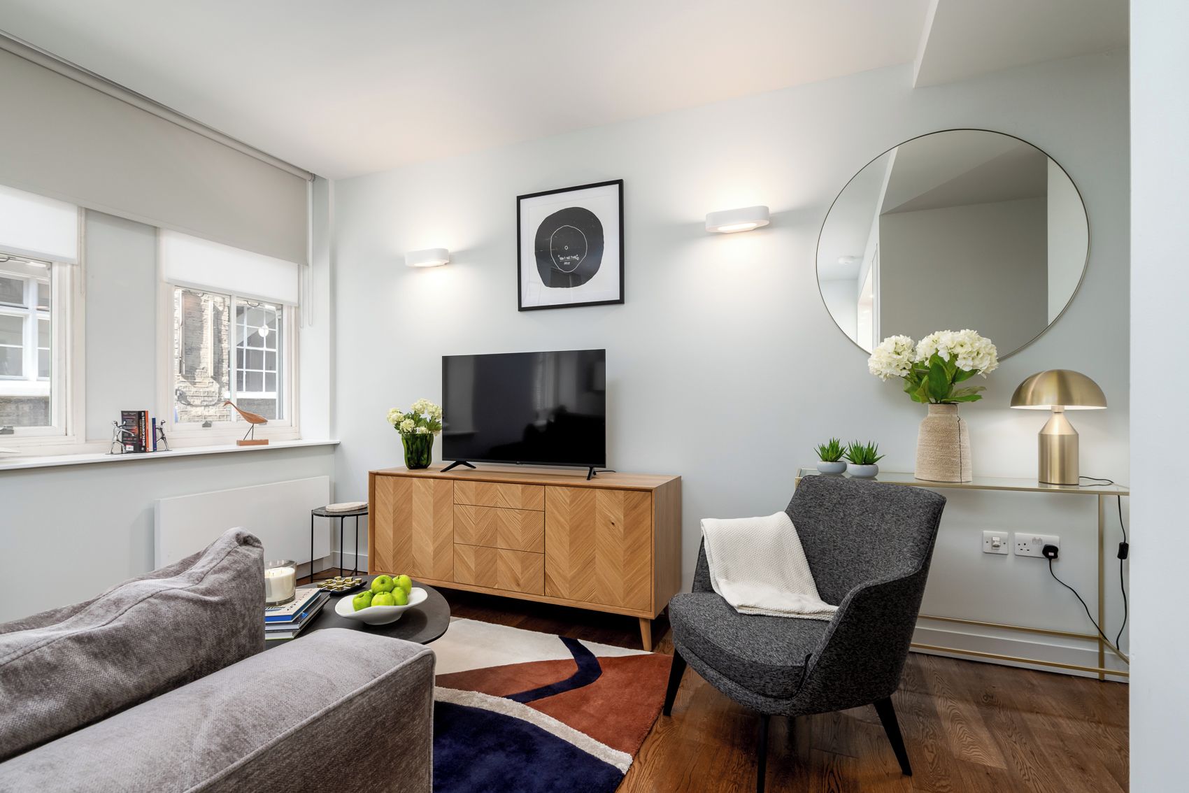 Ein modernes Wohnzimmer mit Flachbildfernseher, Holzschrank, grauem Sessel und rundem Spiegel an der Wand.
