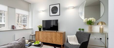 Ein modernes Wohnzimmer mit Flachbildfernseher, Holzschrank, grauem Sessel und rundem Spiegel an der Wand.