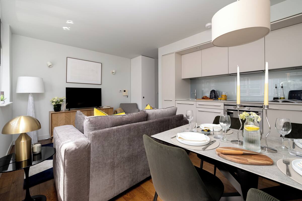 A modern living room with a grey sofa, a dining table set for two, and a kitchen area with white cabinets.