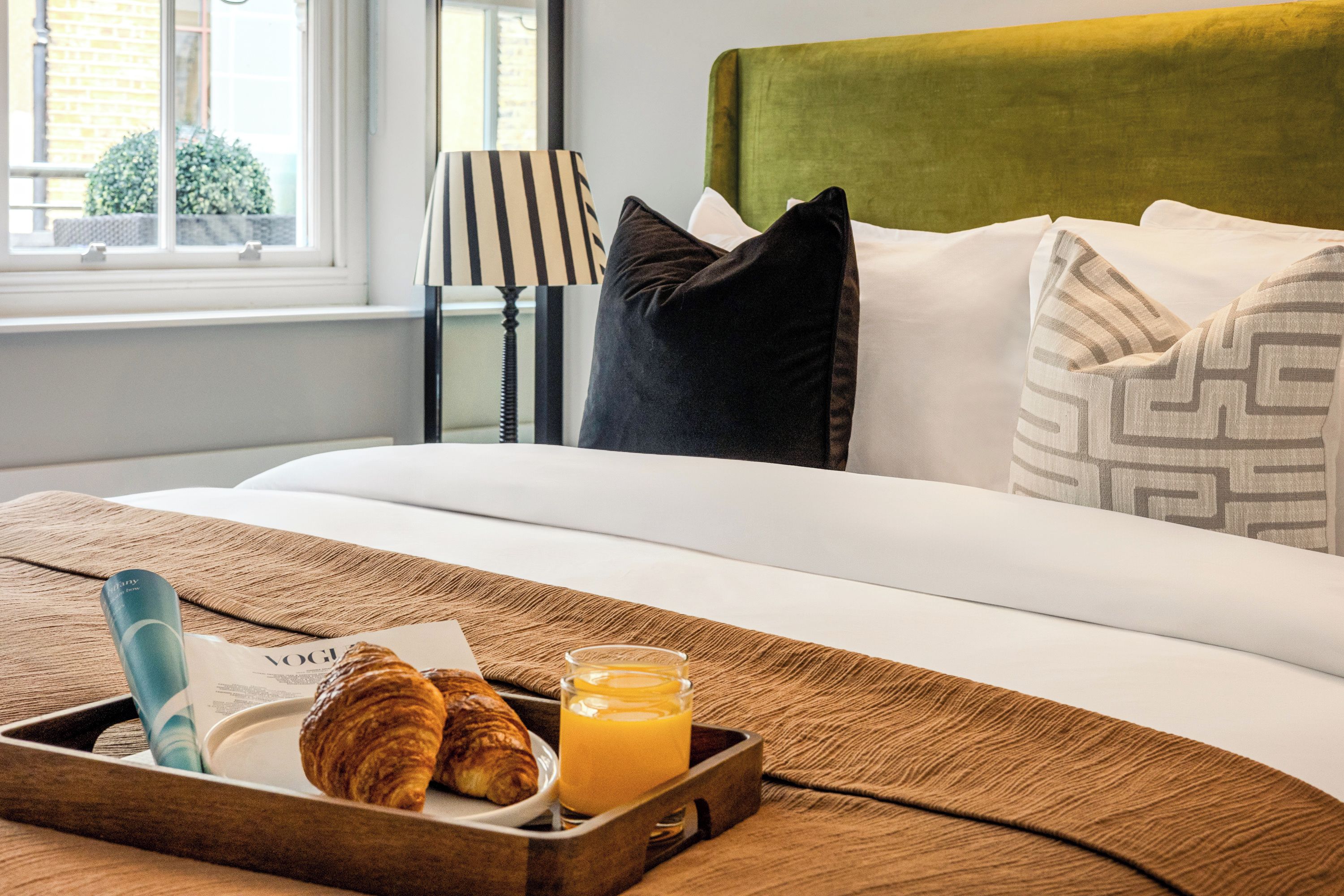 A neatly made bed with a green headboard, a bedside lamp, and a tray with croissants and orange juice.