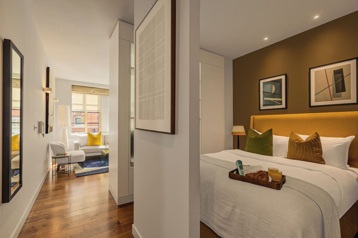A hotel room with a large bed, a bedside table with a tray of breakfast items, a window with a view, and a hallway leading to another room.
