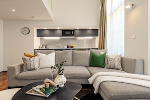 A modern living room with a grey sectional sofa, a coffee table with books and a vase, and a kitchen area with a microwave and cabinets in the background. - Liverpool Street - Your Apartment (London)