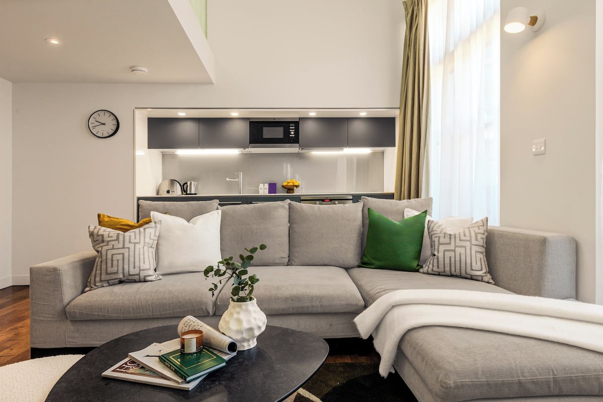 A modern living room with a grey sectional sofa, a coffee table with books and a vase, and a kitchen area with a microwave and cabinets in the background.