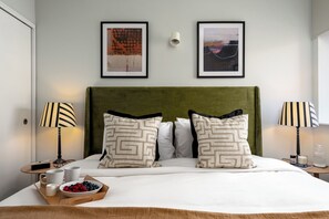 A neatly made bed with a green headboard, two bedside tables with lamps, and two framed artworks on the wall. - Liverpool Street - Your Apartment (London)