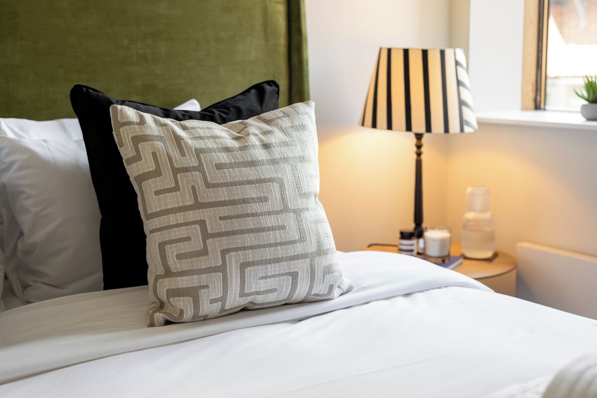 A neatly made bed with white bedding, a geometric patterned pillow, and a striped lamp on a bedside table.