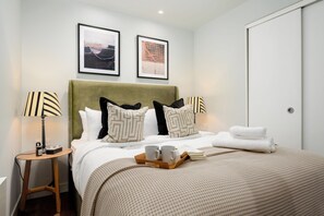 A neatly arranged hotel room with a bed, two bedside tables, a nightstand, a tray with cups and a book, and two framed pictures on the wall. - Liverpool Street - Your Apartment (London)