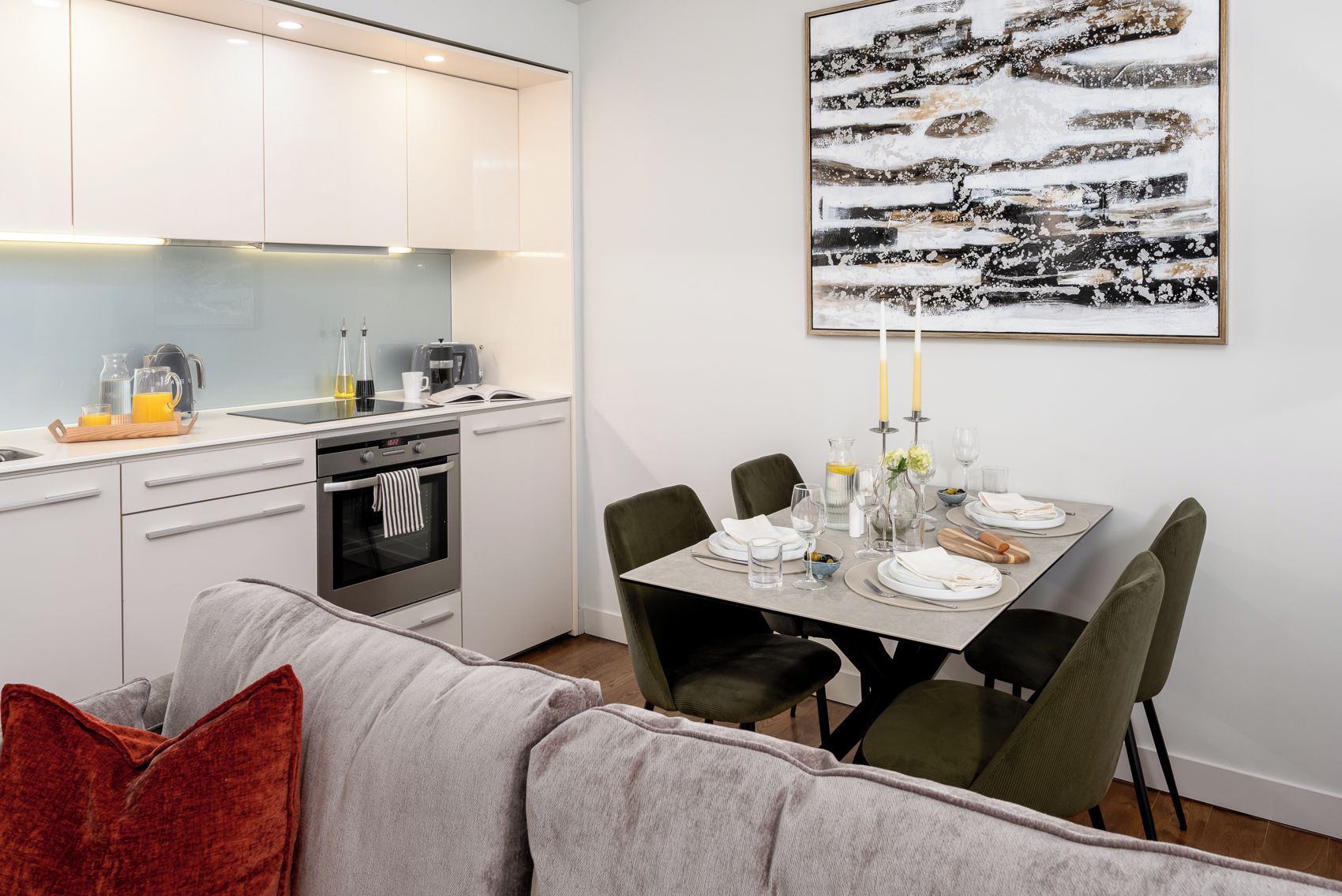 Una cucina moderna con mobili bianchi, un tavolo da pranzo per quattro persone e un grande quadro astratto appeso al muro.