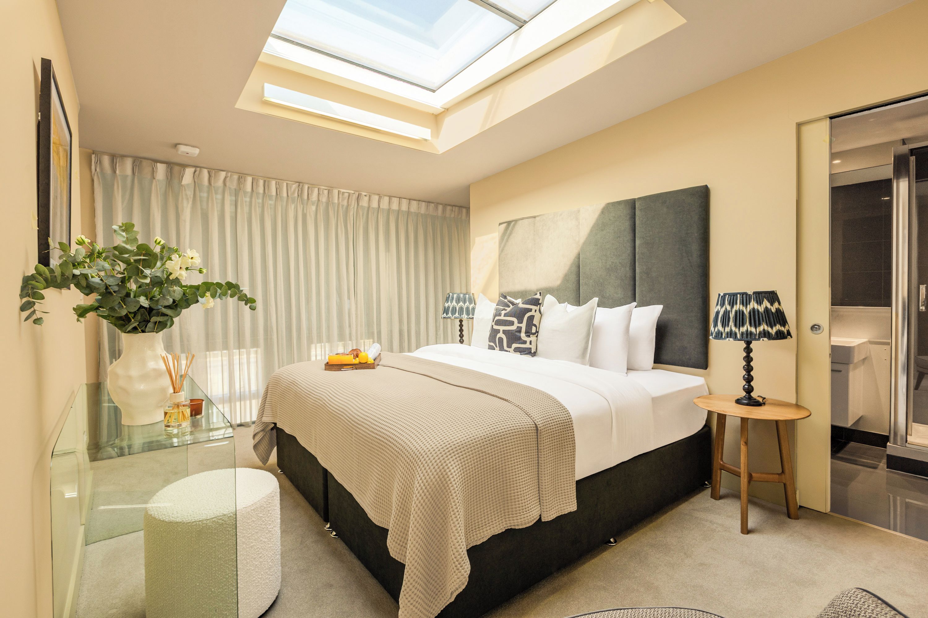Ein modernes Schlafzimmer mit Dachfenster, einem Glastisch, einem Nachttisch und einem großen Bett mit Kopfteil.