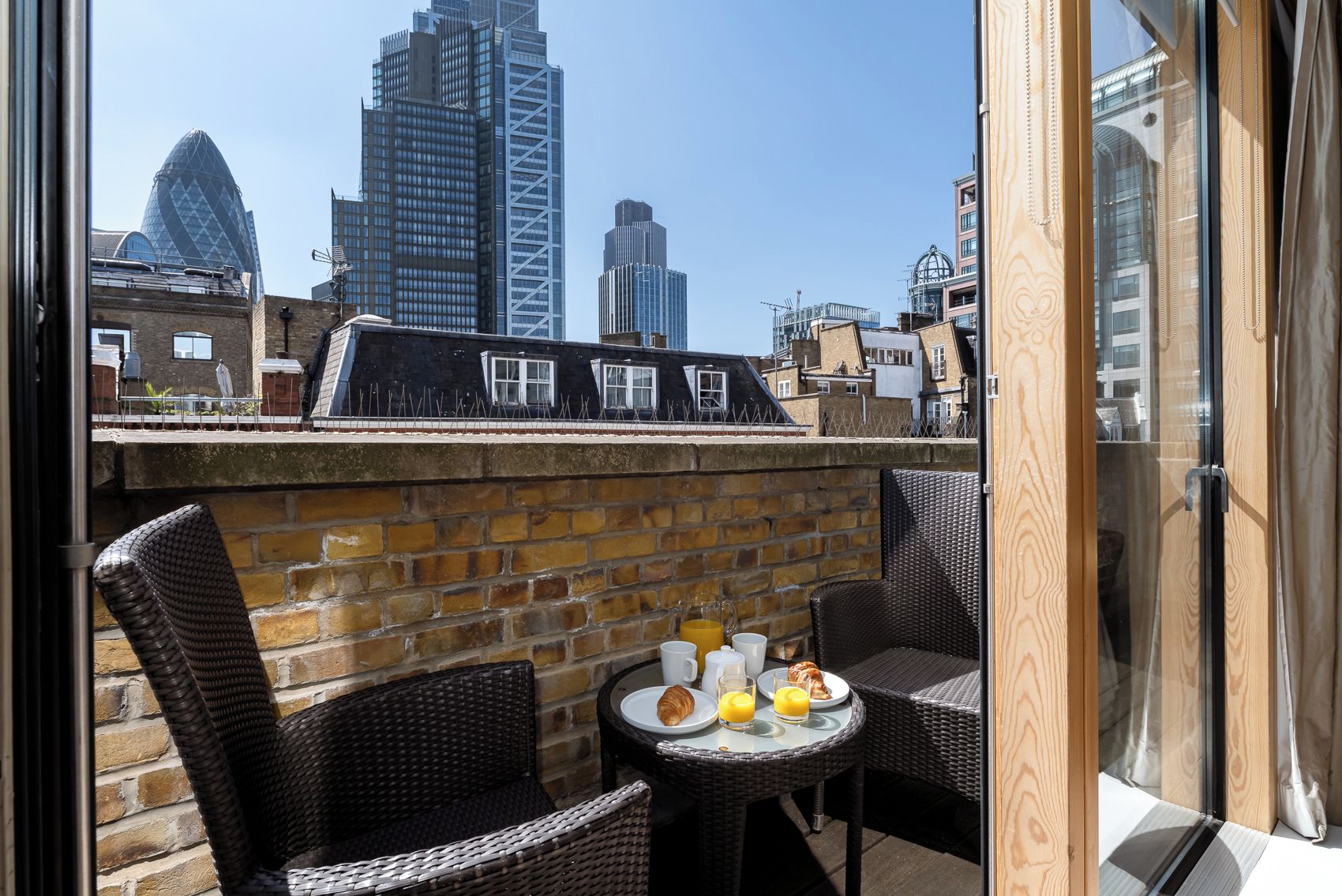 A rooftop terrace with wicker furniture, a brick wall, and a table set for breakfast.