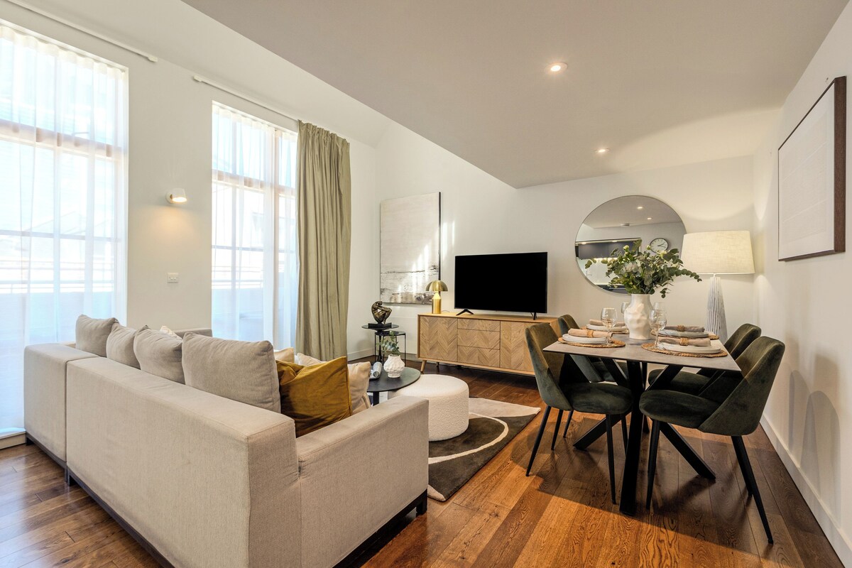 A modern living room with a sofa, dining table, and chairs. There is a flat-screen TV, a mirror, and a window with curtains.