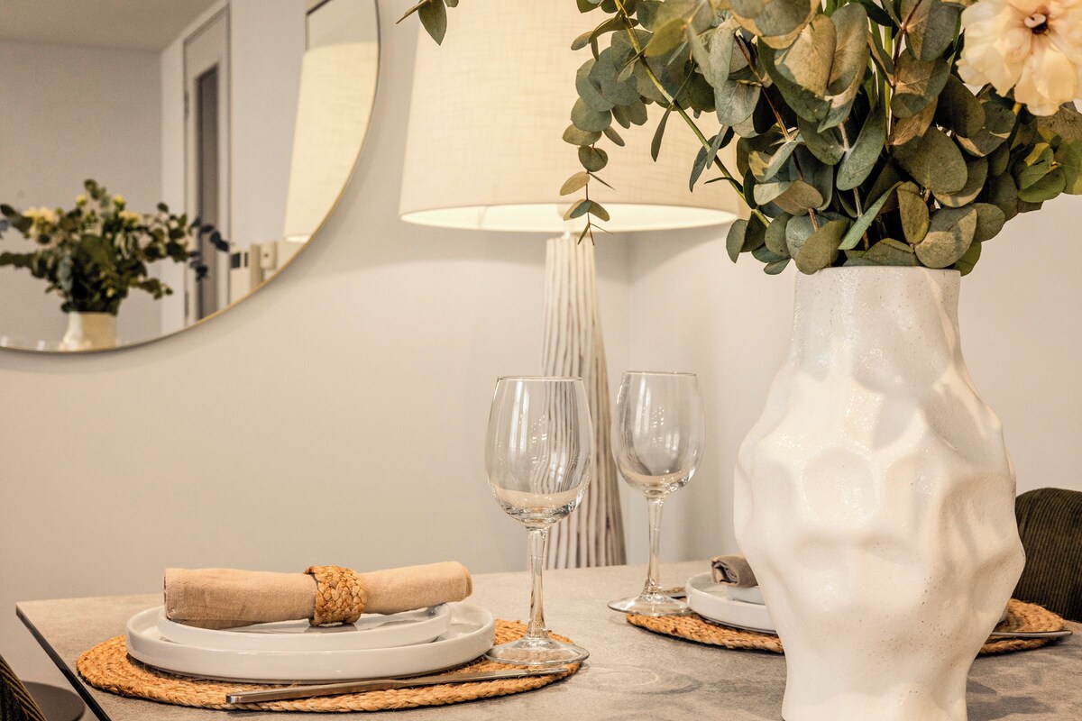 A dining table with a white vase, two wine glasses, and a rolled napkin.