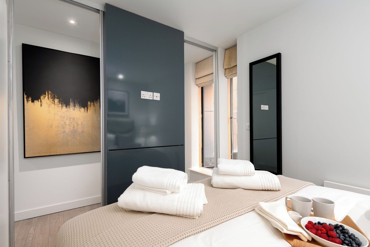 A modern hotel room with a large bed, a painting on the wall, and a breakfast tray with fruits and coffee.