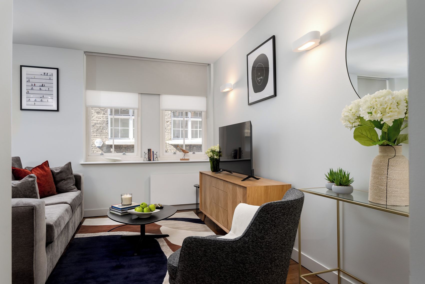 Una sala moderna con un sofá, una mesita de centro con libros y manzanas, un televisor sobre un mueble de madera y un florero con flores.