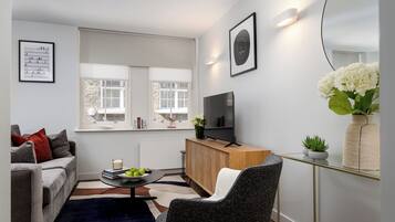 Una sala moderna con un sofá, una mesita de centro con libros y manzanas, un televisor sobre un mueble de madera y un florero con flores.