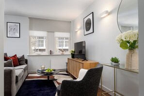 A modern living room with a sofa, a coffee table with books and apples, a TV on a wooden stand, and a vase with flowers. - Liverpool Street - Your Apartment (London)