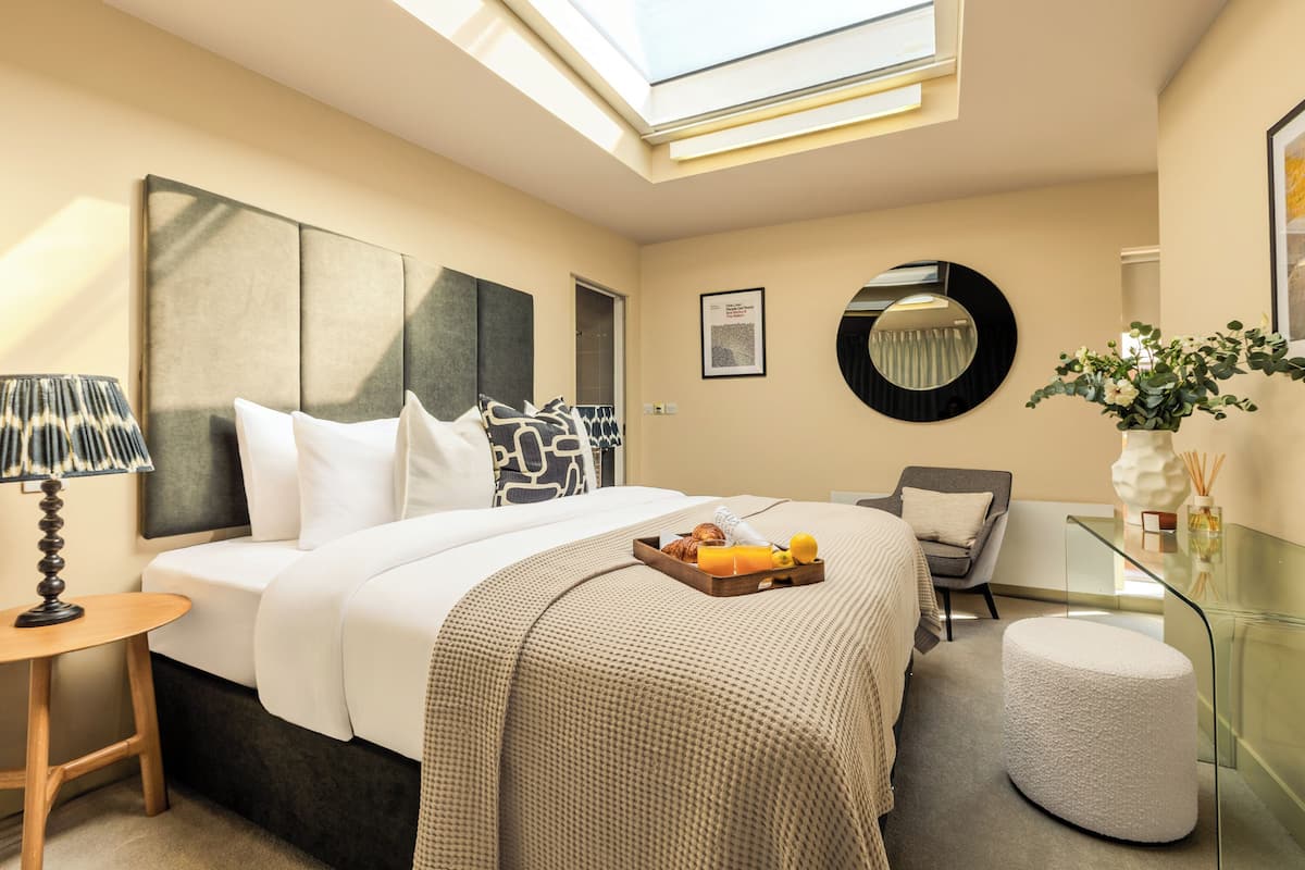 A modern hotel room with a large bed, a glass coffee table, a round mirror, and a small round ottoman.