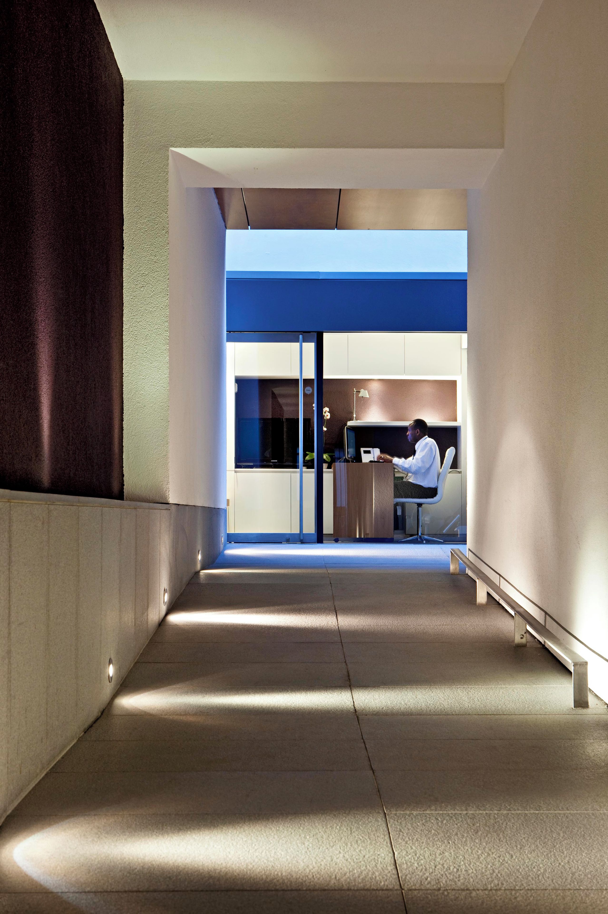 A modern hallway with a person at a desk, a glass door leading to a room, and a bench along the wall.