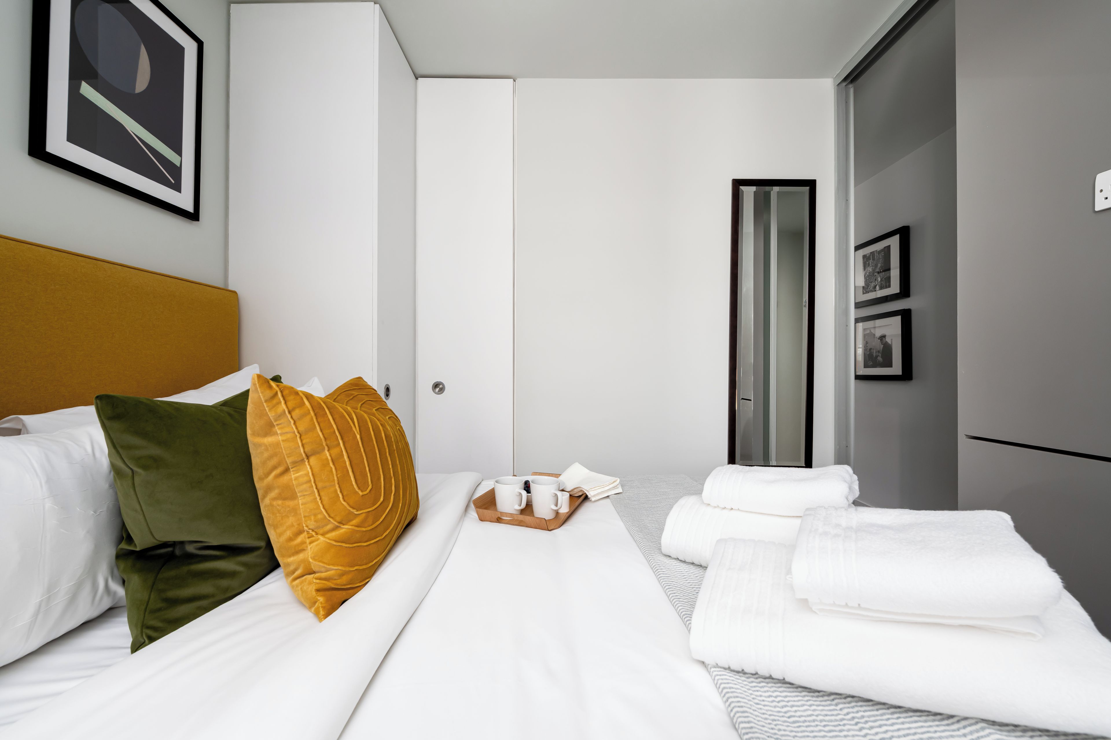 A hotel room with a bed, a bedside table with a tray of white cups and a book, and a wardrobe.