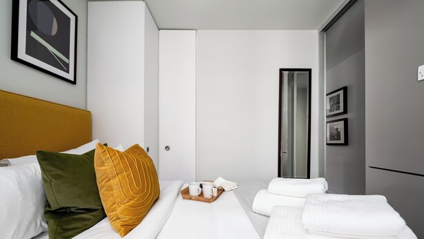 A hotel room with a bed, a bedside table with a tray of white cups and a book, and a wardrobe. - Liverpool Street - Your Apartment (London)